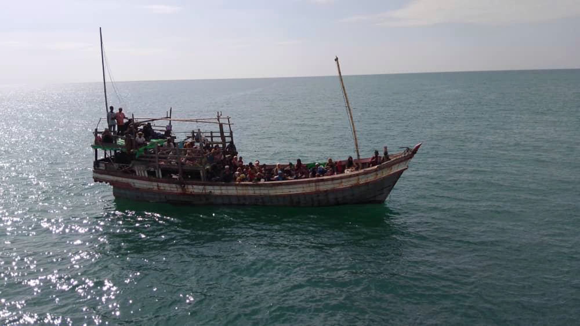 A wooden boat crowded with people sails on a calm sea under a clear sky.