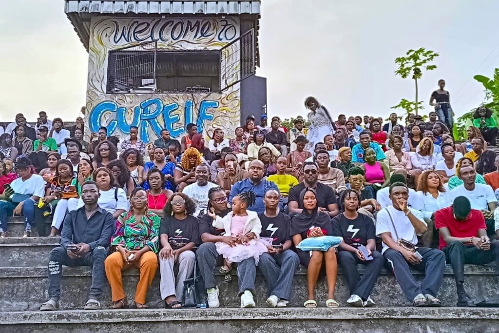 A large group of people sit on outdoor steps, with a sign behind them reading "Welcome to Cherefe."