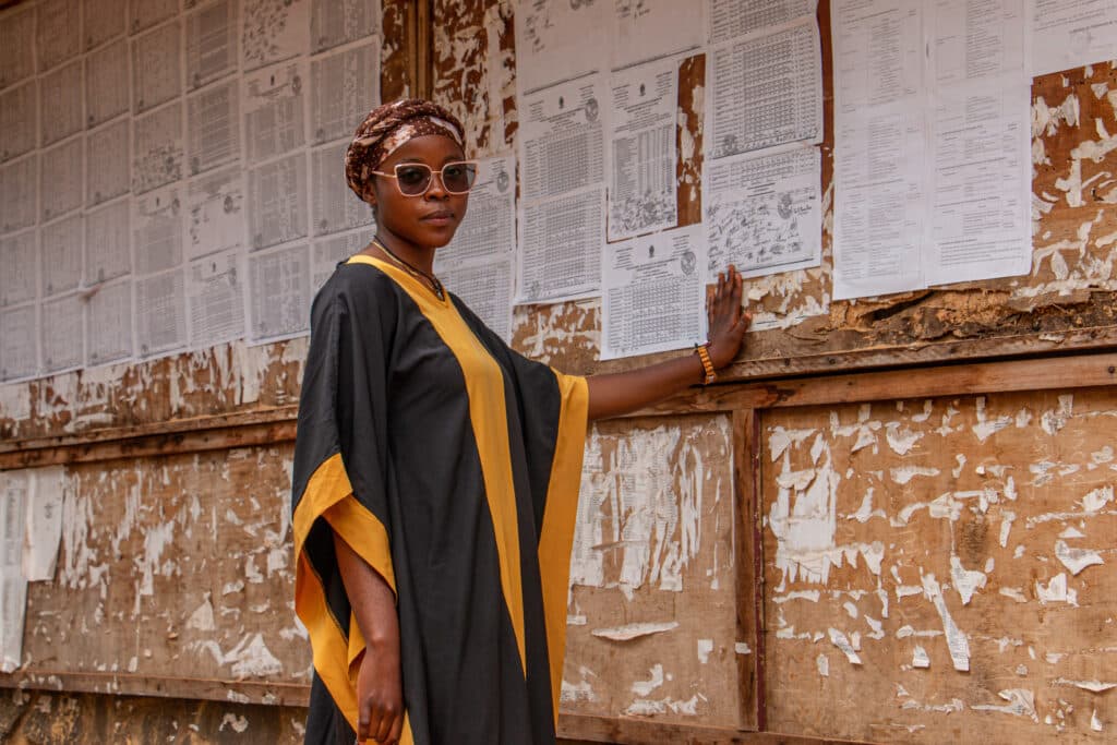 A woman in sunglasses and a black and yellow robe stands against a weathered wall covered in papers.