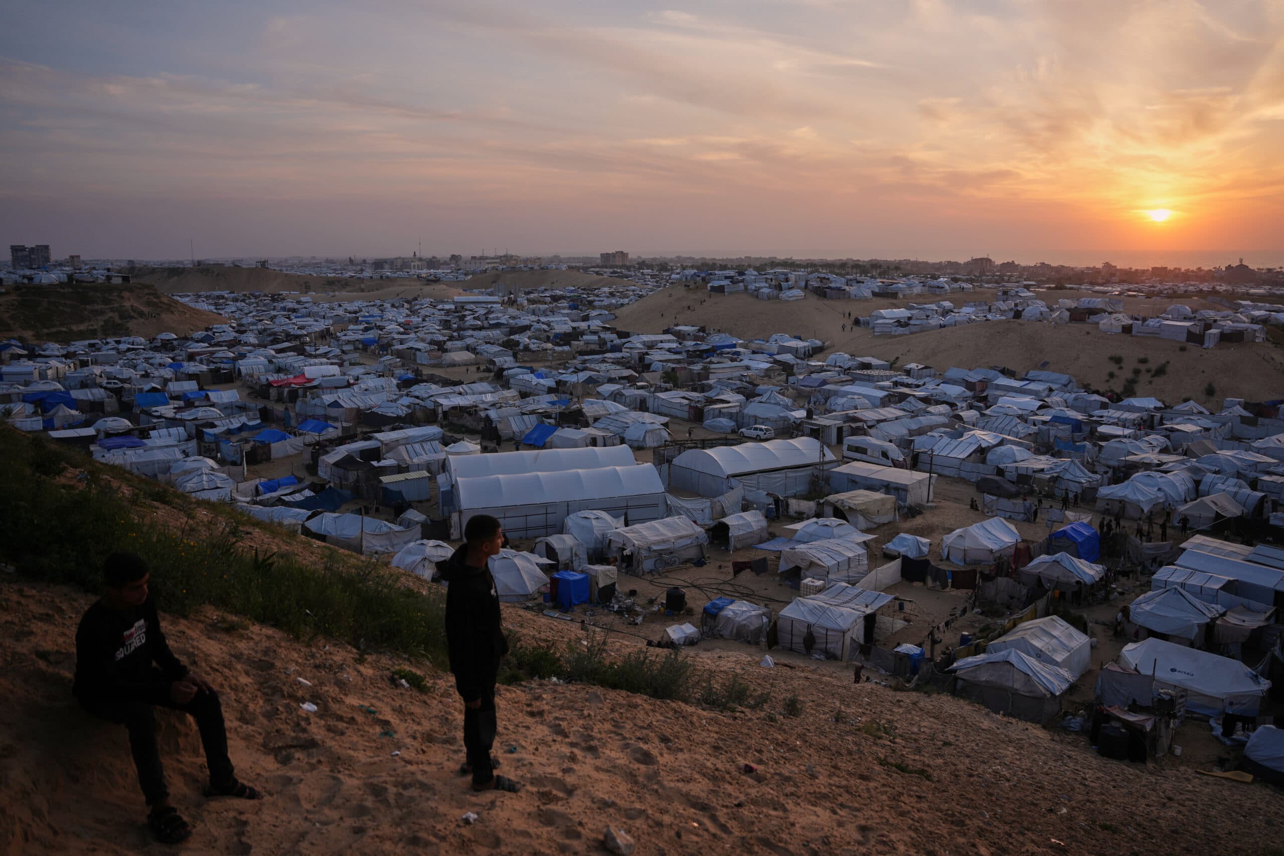 Two people stand on a hill overlooking a large refugee camp at sunset, with numerous tents and makeshift shelters visible.