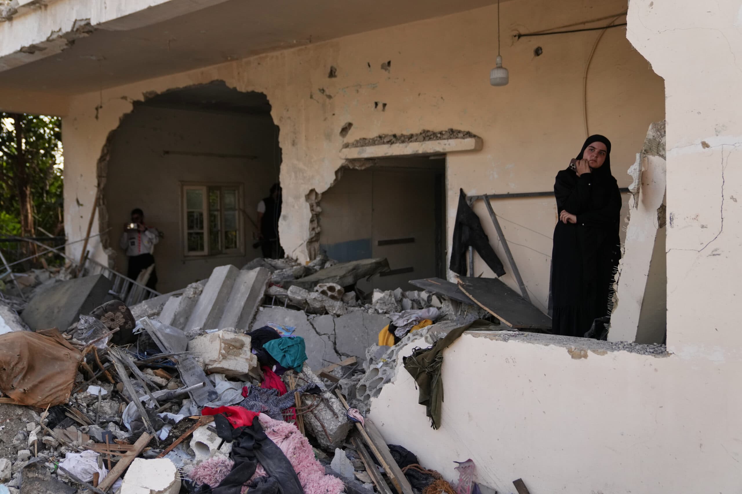 A woman in dark clothing stands amid rubble inside a partially destroyed building with a large hole in the wall.