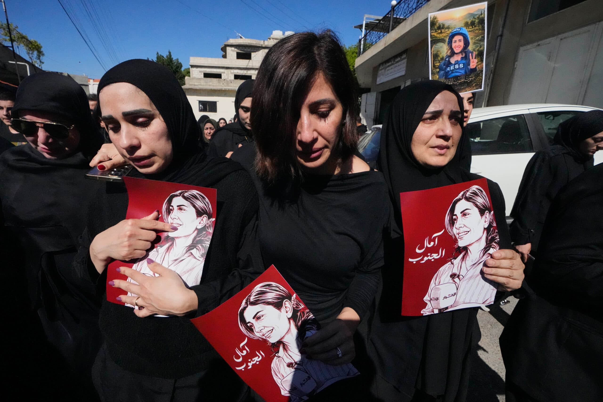 A group of women in black clothing hold illustrated posters of a young woman during a solemn outdoor gathering.