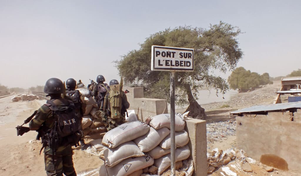 Soldiers in tactical gear stand near sandbags and a sign reading "Pont sur l'Elbeid," with a tree and dry landscape in the background.