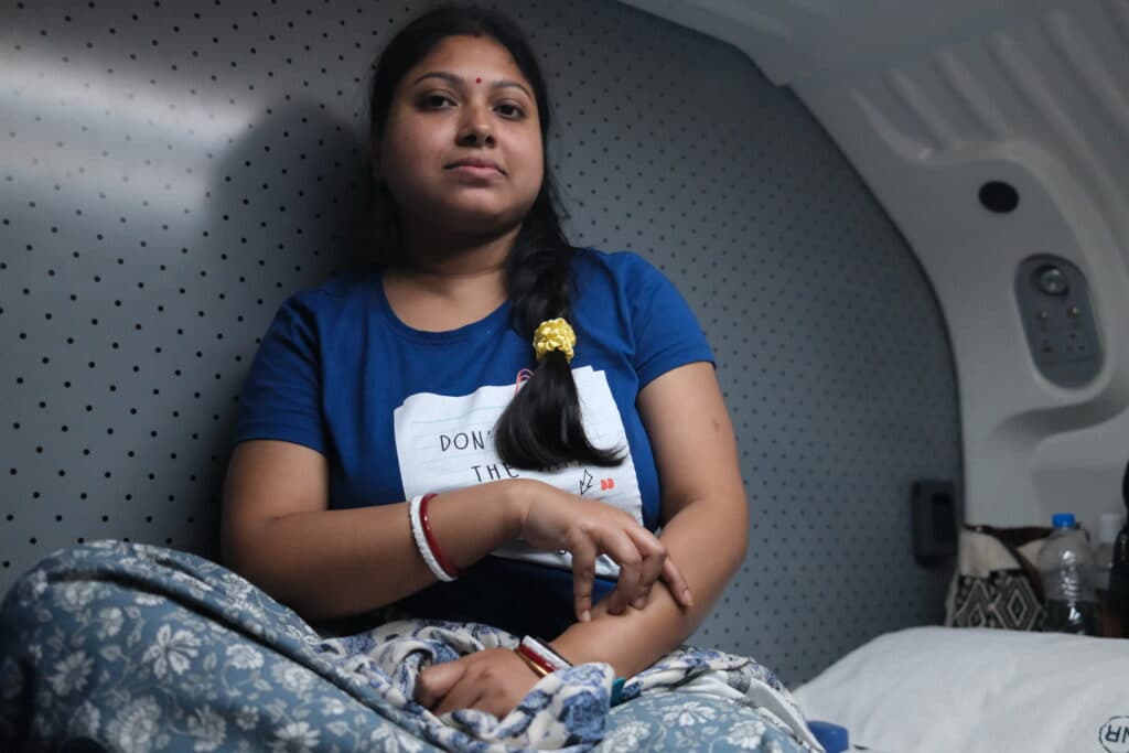 A woman in a blue shirt sits in a vehicle interior, wearing patterned pants with her arm resting on her knee.