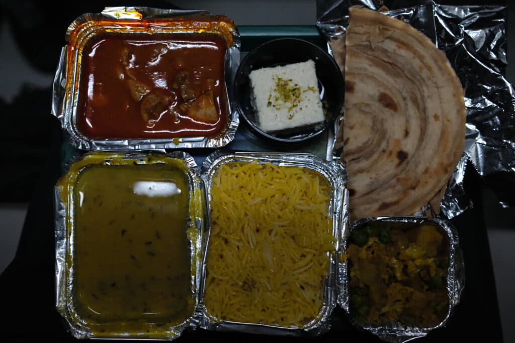 A meal tray with various Indian dishes, including dal, rice, curry, a dessert square, and a paratha.