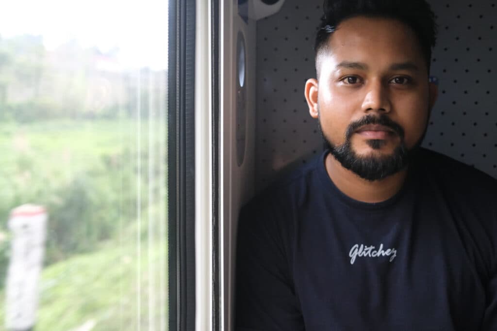 A man with a beard sits by a window on a train, wearing a shirt with "Glitcher" logo.