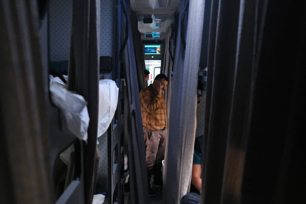 A person standing in a narrow corridor between bunk beds on a train, talking on a phone.