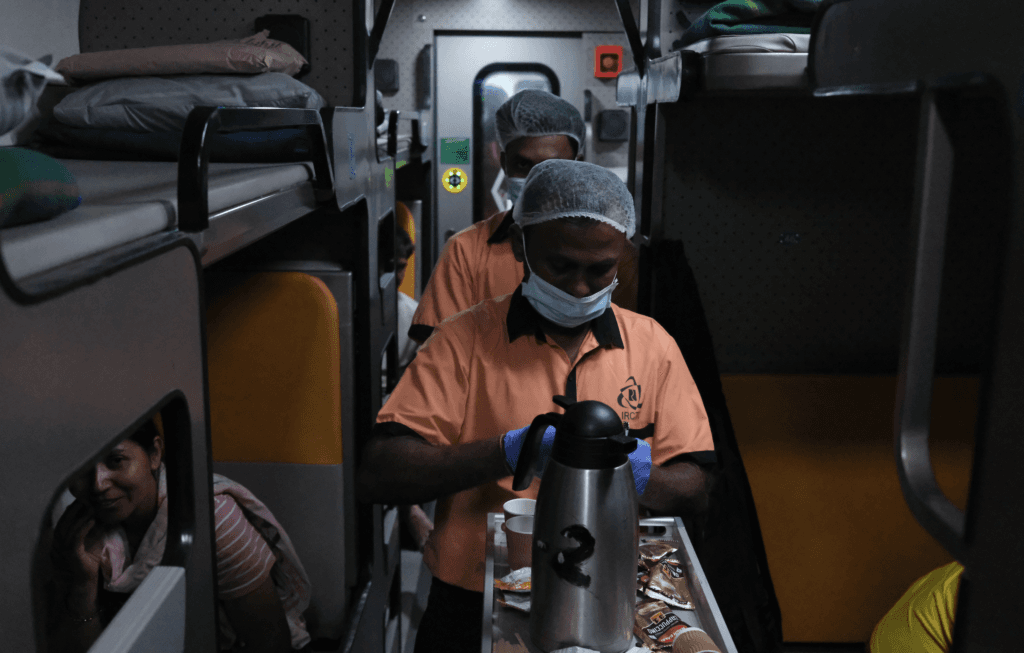 Two staff members in uniforms serve beverages from a trolley in a train compartment, with bunks visible.