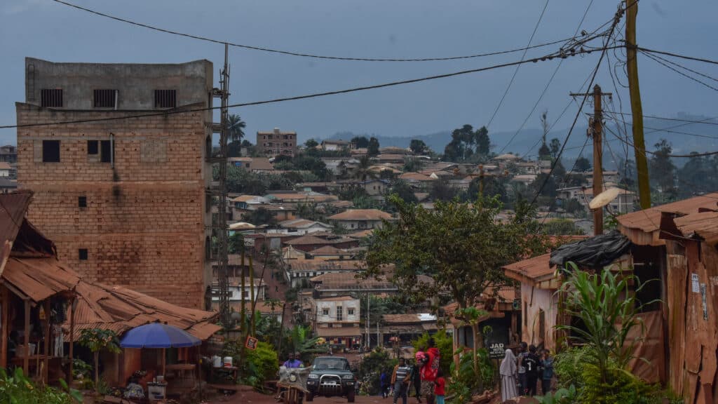 A busy street in a hilly town with people walking, shops, utility poles, and a backdrop of trees and houses under a cloudy sky.
