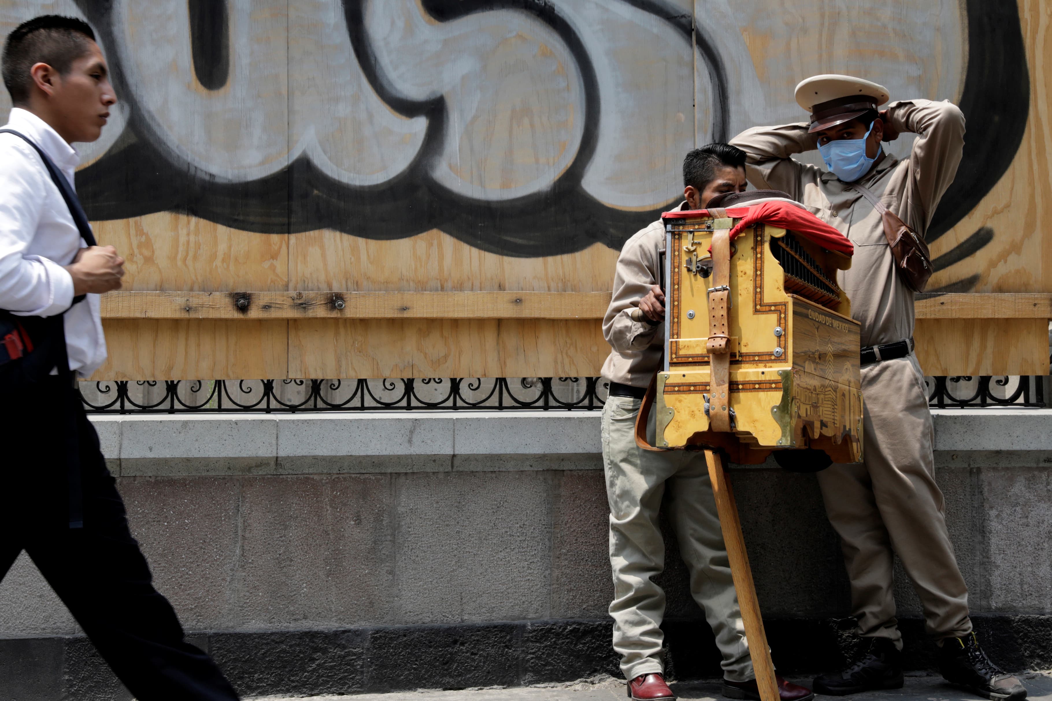 An organ grinder wears a surgical mask on a sidewalk as a man walks past.
