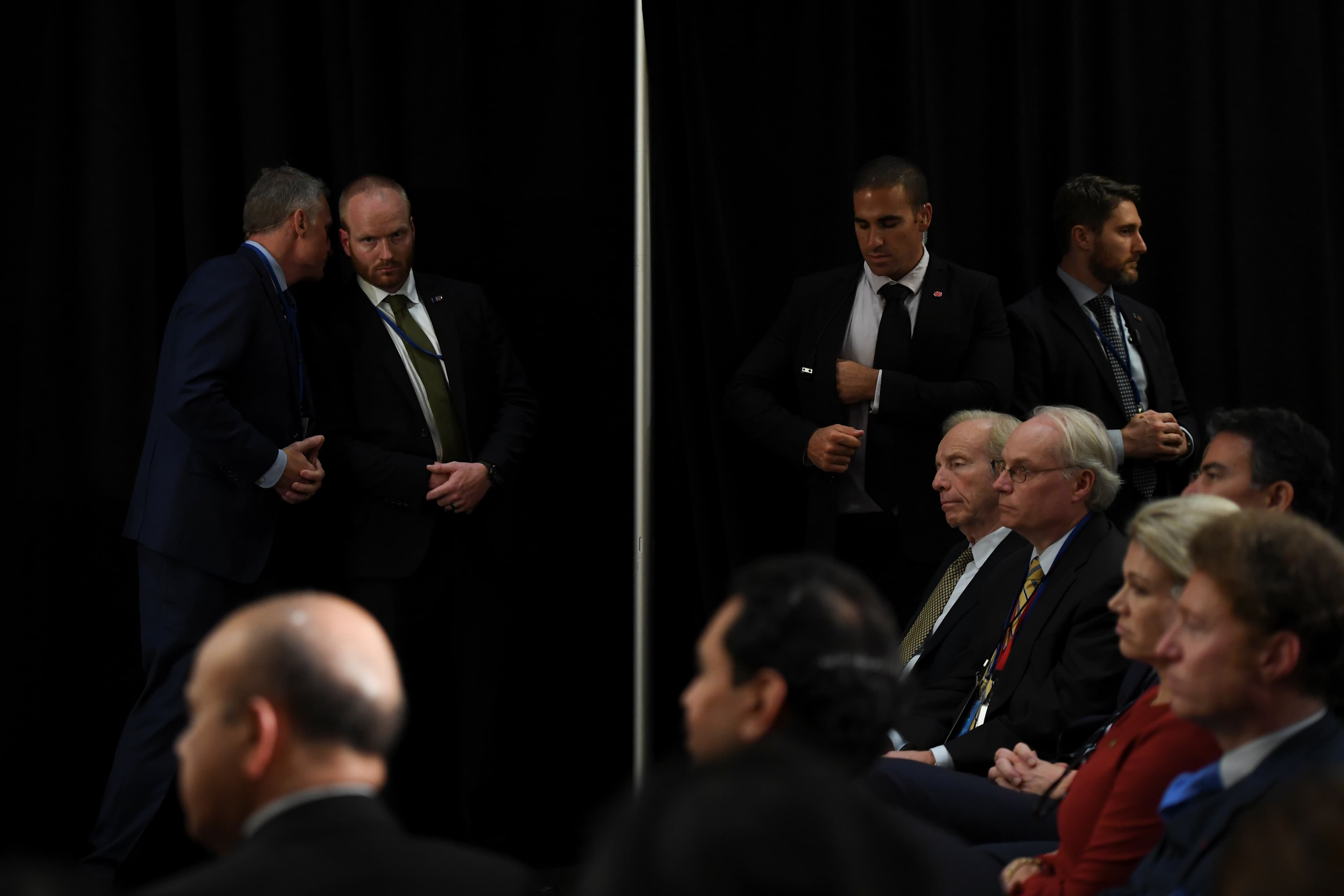 Several men in dark suits stand as others sit