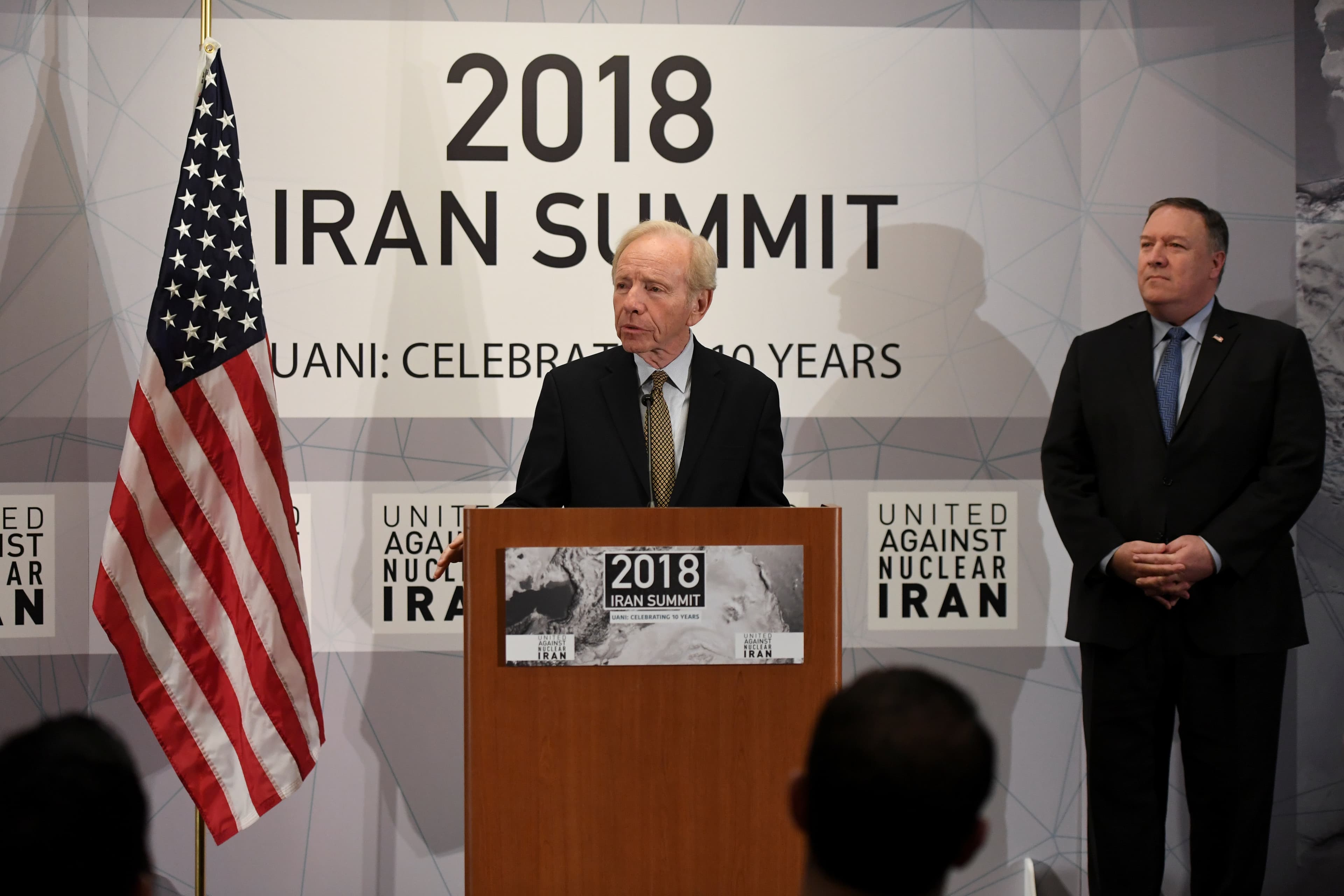 A man speaks at a podium with another man standing near him and the American flag to one side