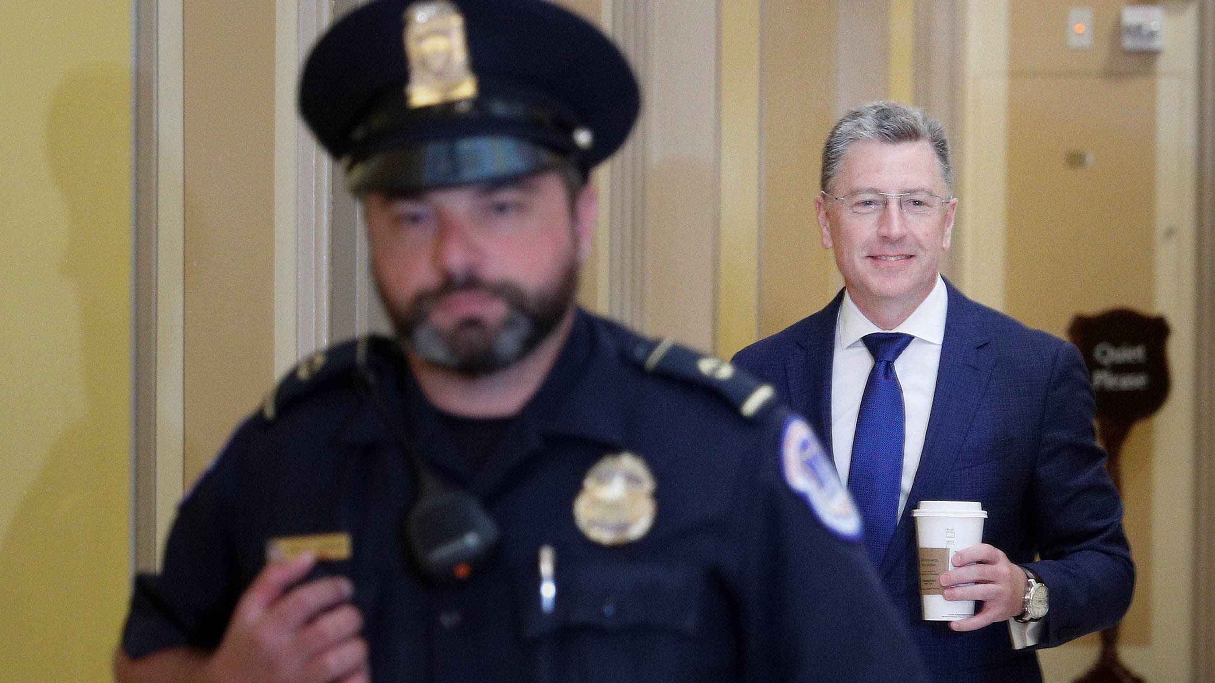 A uniformed officer walks in front of a man in a suit smiling and holding a coffee