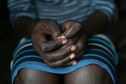 A woman sits with her hands folded on her lap, wearing a blue dress.