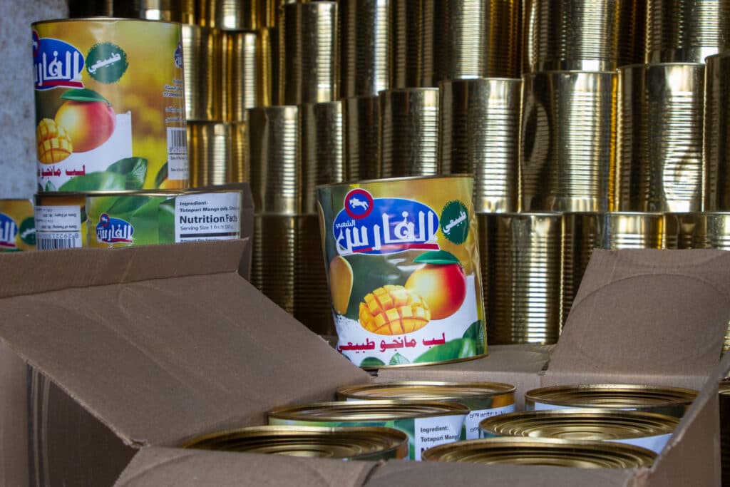 Cans of mango pulp with colorful labels in a cardboard box, with a stack of plain metal cans in the background.
