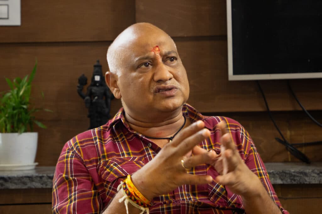 A man in a checkered shirt with a tilak on his forehead, gesturing with his hands, sits indoors with a small plant and a statue in the background.