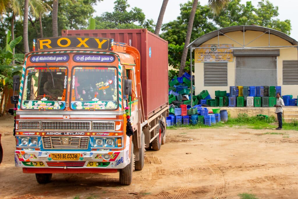 A colorful Indian truck labeled "ROXY" parked in front of a building with stacks of plastic crates, surrounded by green trees and a man standing nearby.