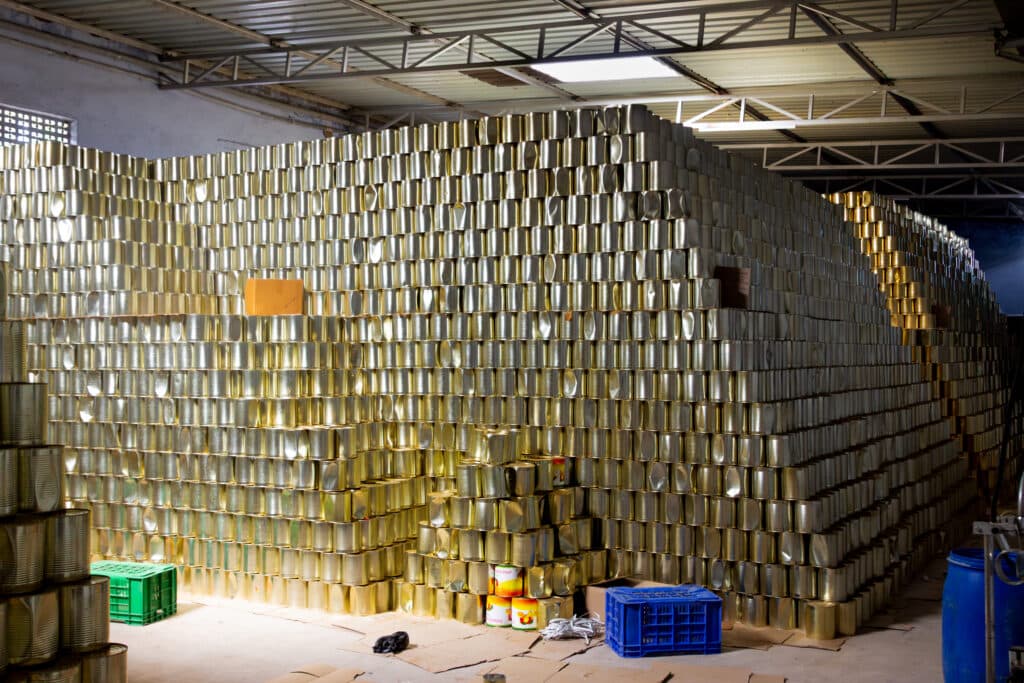 A large stack of empty metal cans organized in a storage area, with a few colored crates and a blue barrel nearby.