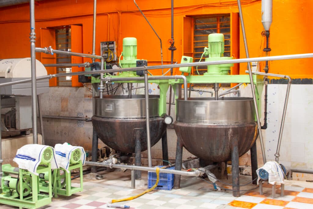 Industrial kitchen setting with two large metal mixing vats equipped with green motors and piping, against an orange and tiled wall.