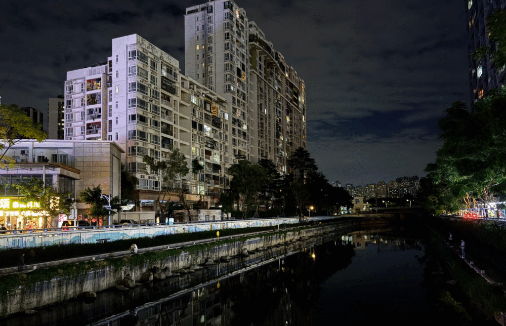 A nighttime view of a canal flanked by high-rise apartment buildings in an urban setting. The buildings are illuminated, reflecting in the calm water of the canal, while nearby trees are lightly lit. The sky is cloudy with faint ambient lighting from the city.