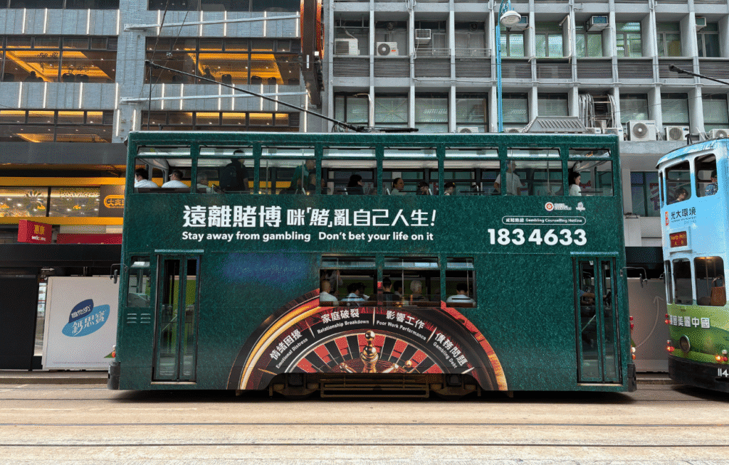 A green double-decker tram featuring an anti-gambling advertisement with the text "Stay away from gambling. Don’t bet your life on it," and the hotline number 1834633. The side of the tram displays a roulette wheel design and various consequences of gambling like "Relationship Breakdown" and "Poor Work Performance." The tram is stopped near a street with commercial buildings in the background.