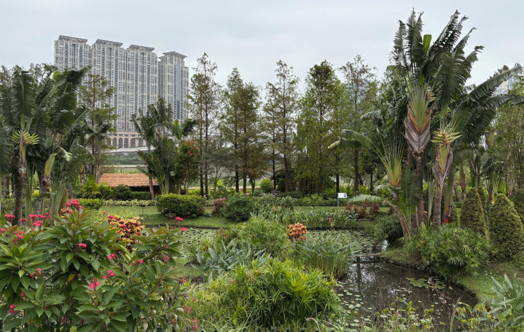 A lush garden with various tropical plants and a small pond, bordered by a cluster of tall trees. In the background, a tall, modern apartment building is visible under an overcast sky.