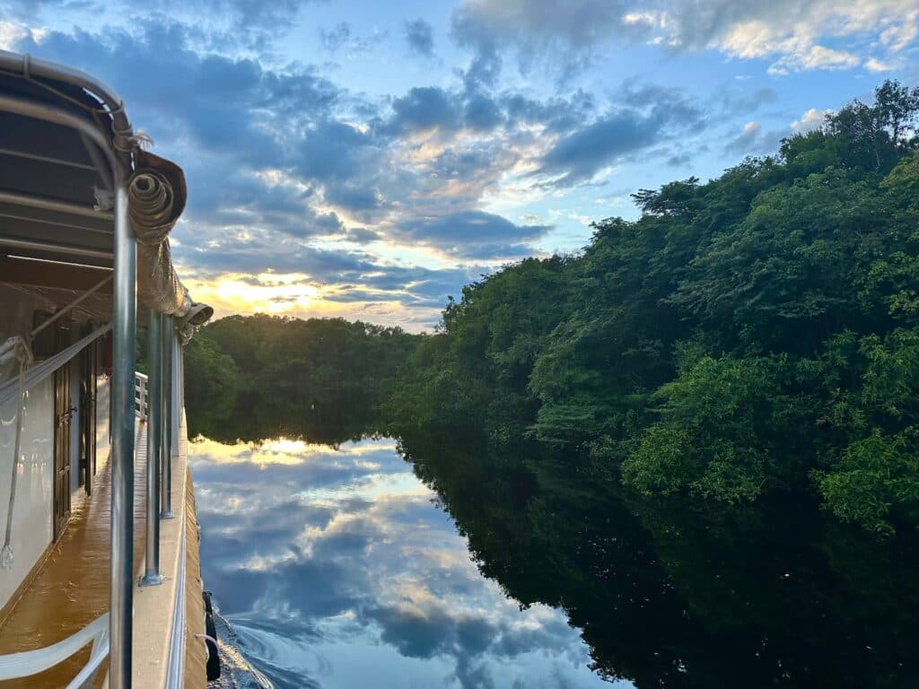 A boat travels down a calm river surrounded by dense, lush green trees, with the sky reflecting beautifully on the water as the sun sets in the background.