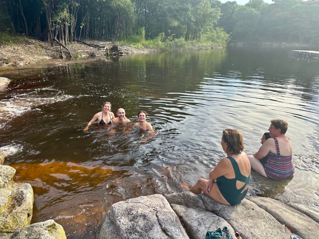 Three people are swimming in a natural pond while two others sit on rocks at the water's edge. The surrounding area is lush with green trees.