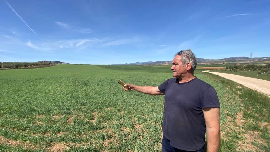A man standing in a field, holding a smartphone, with a dirt path and rolling hills in the background under a clear blue sky.
