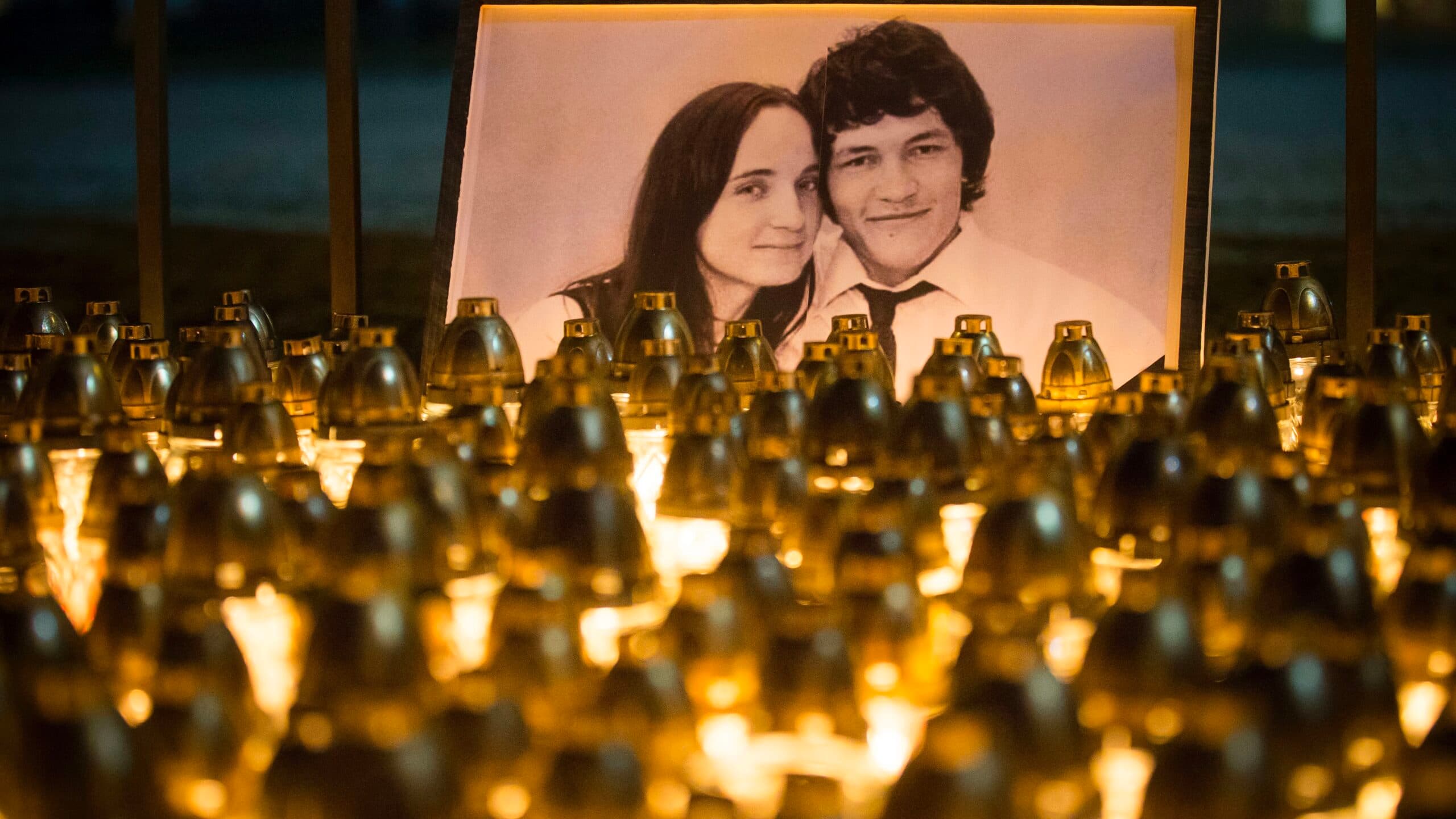 A photograph of a couple is displayed alongside numerous lit candles, creating a memorial scene with warm, glowing light.