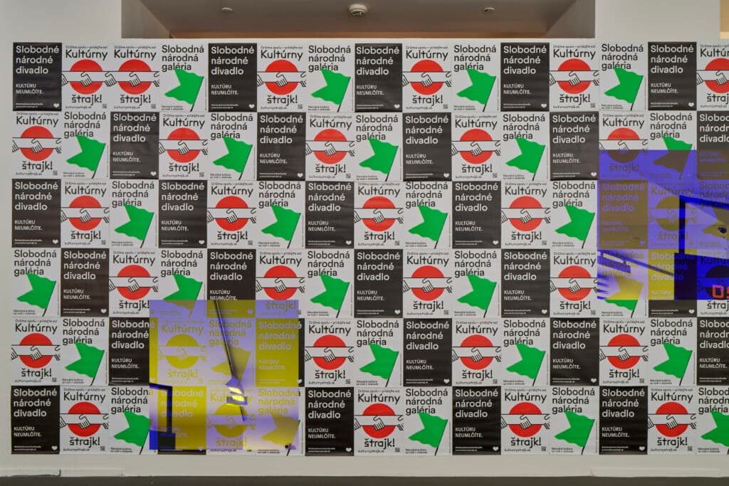 A wall covered with posters featuring Slovak text and graphic designs, including a red circle with a horizontal line and a green arrow shape. The posters have repetitive text and color variations, such as black, white, red, green, and yellow, creating a striking pattern.