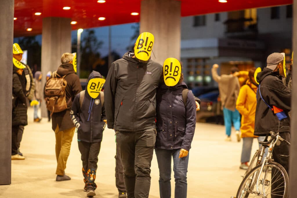 A group of people wearing yellow face masks with a frowning expression and the letters 'B!B' on them, gathered in an outdoor area with a red ceiling, some carrying backpacks and a bicycle visible on the right.