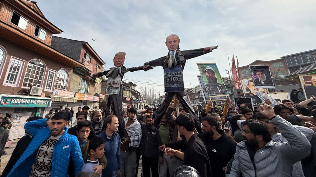 A large crowd of people gather on a street, holding up effigies and signs during a protest. The effigies depict political figures, and the crowd appears engaged in a demonstration, with some holding portraits of other figures. The scene is set in a bustling urban area with a backdrop of traditional buildings and shops.