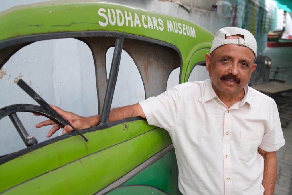 A man wearing a white shirt and a backward cap stands next to a green vehicle at Sudha Cars Museum.