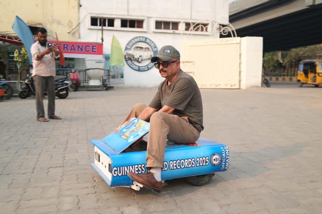A man wearing sunglasses and a cap is sitting on a small, novelty vehicle resembling a miniature race car, marked with 'Guinness World Records 2025'. Another man in the background is taking a photo with a phone.