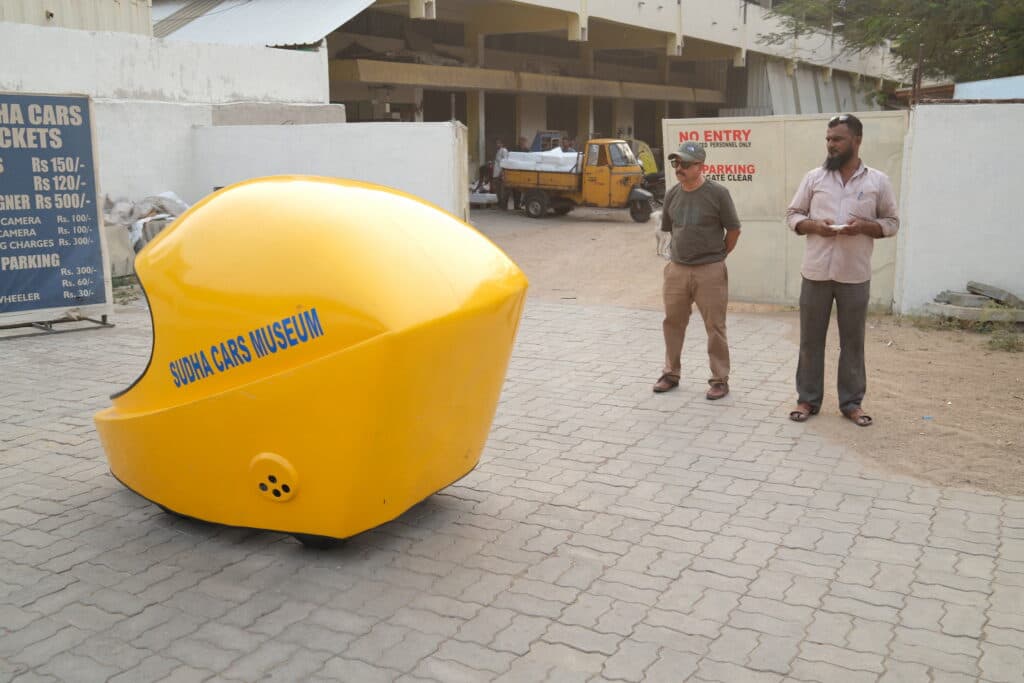 A yellow, helmet-shaped vehicle is parked in front of the Sudha Cars Museum. Two men stand nearby observing it, with one wearing a cap and the other holding a small item. Behind them are signs displaying museum ticket information and entry restrictions. The background includes a partially visible building with an open area.